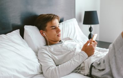 16 years old teenager using smartphone and listening to music in earphones while relaxing on couch in his room